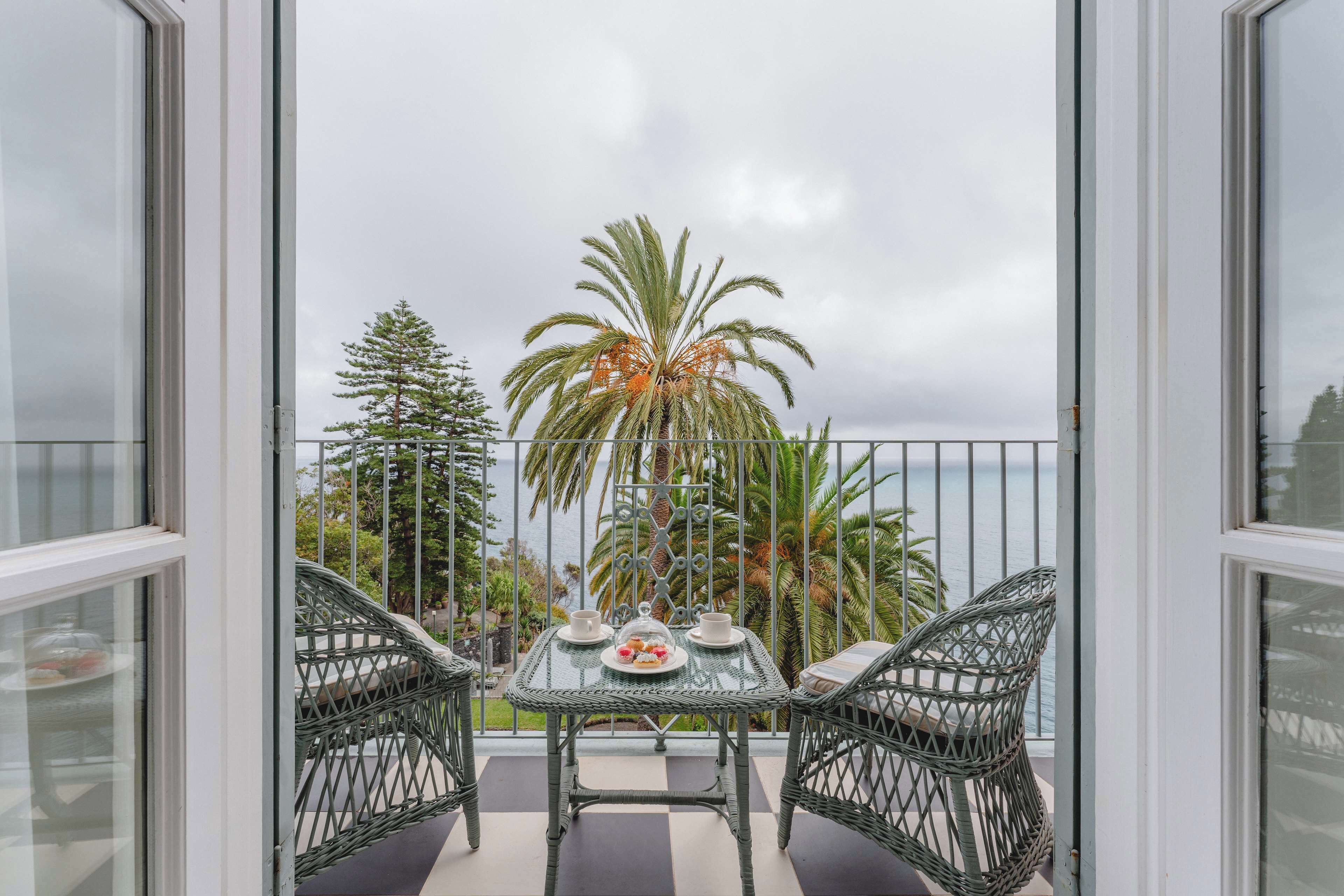 A balcony with two wicker chairs and a table set for two, overlooking a palm tree and the sea.