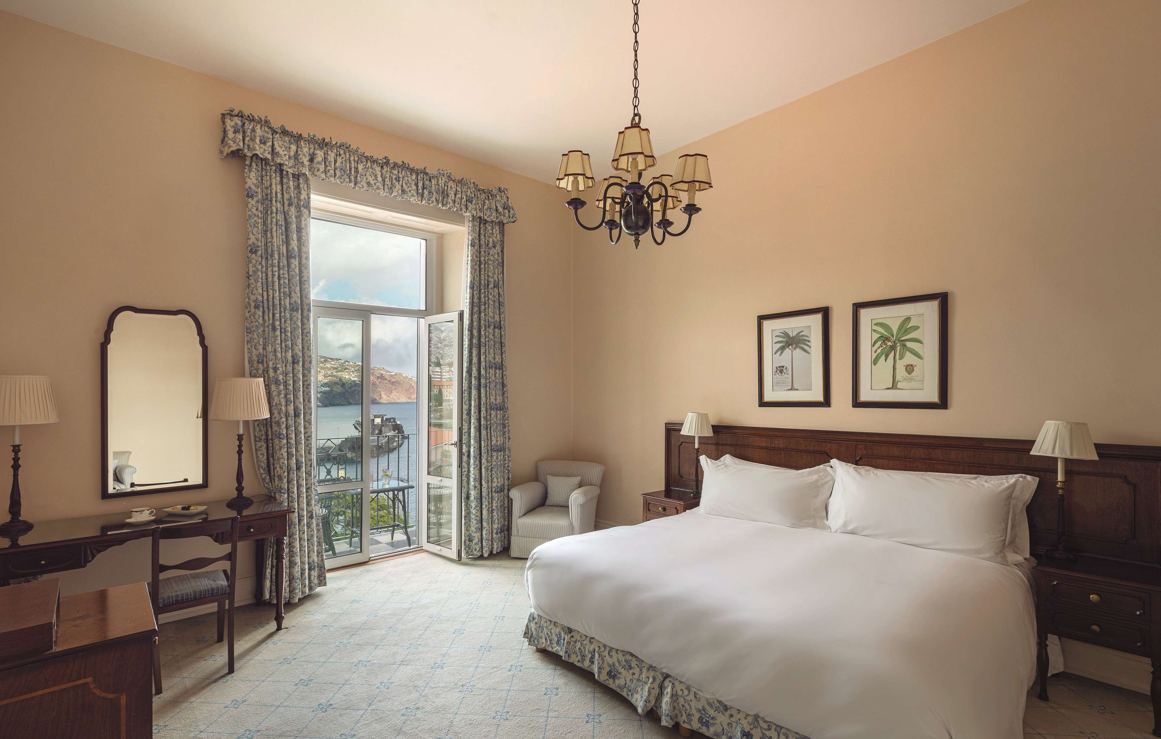 A hotel room with a large bed, a desk, a chair, a nightstand, and a chandelier. There are two framed pictures on the wall and a window with curtains.