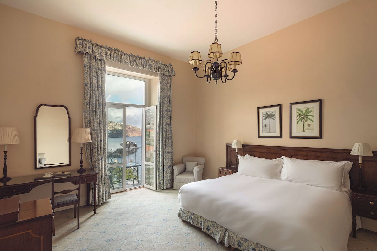 Een hotelkamer met een groot bed, een bureau, een stoel, een nachtkastje en een kroonluchter. Er hangen twee ingekaderde foto’s aan de muur en er is een raam met gordijnen.
