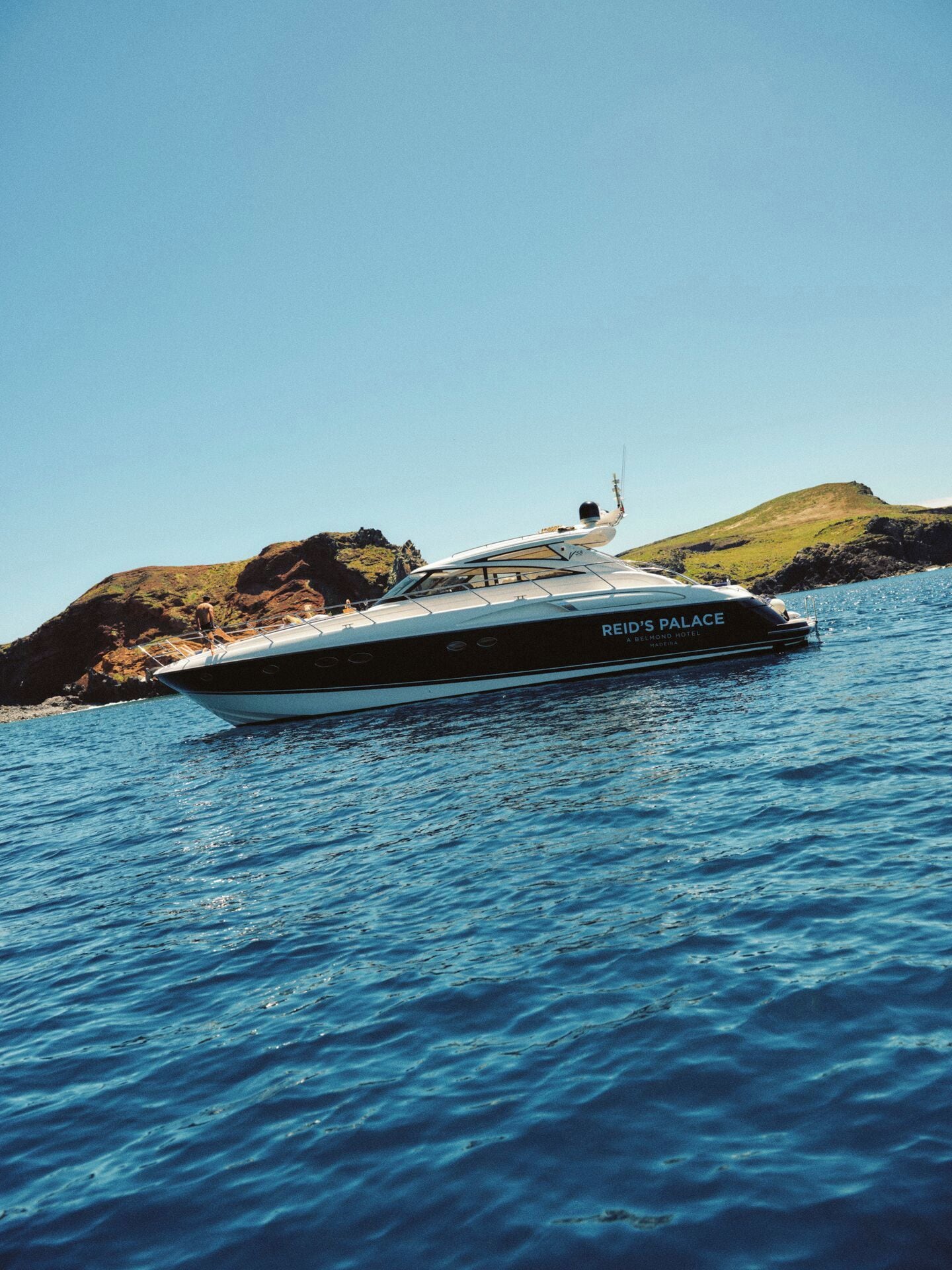 A yacht named 'REID'S PALACE' with a person standing on the deck, surrounded by water and rocky terrain.
