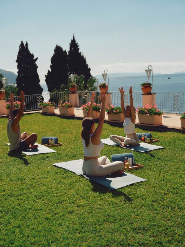 Een groep mensen doet yoga op het gras, met een prachtig uitzicht op zee en bergen op de achtergrond.