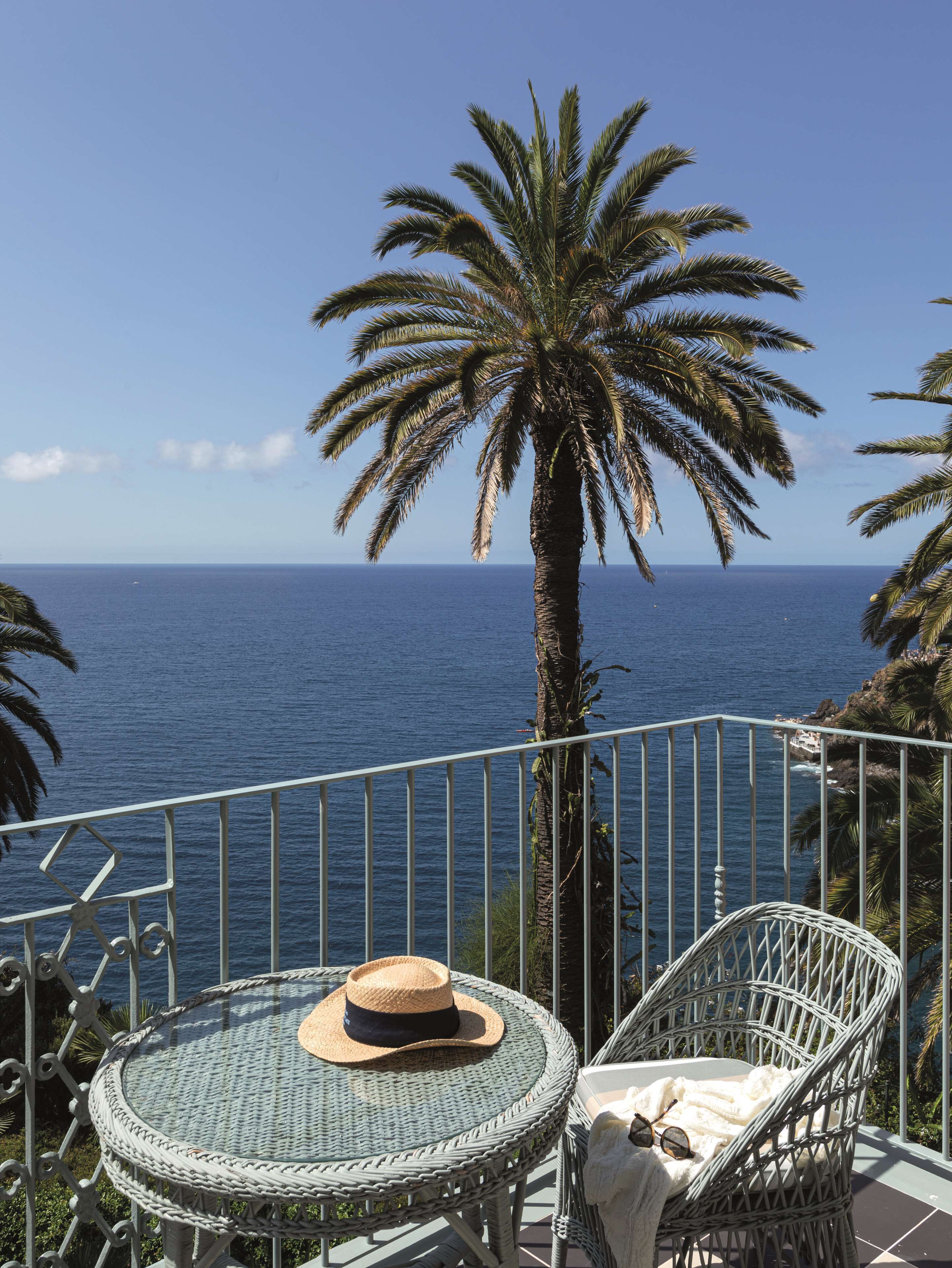 A balcony with a wicker table and chair, a palm tree, and a view of the ocean.