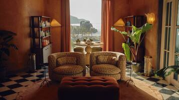 Um quarto com piso xadrez, duas poltronas estampadas, um pufe central, uma mesa redonda com um vaso, uma estante de livros e uma grande janela com vista para a área costeira.