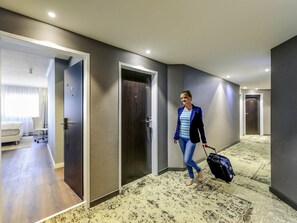 A hotel corridor with doors numbered 505 and 507, a bed in the adjacent room, and a person pulling a suitcase.