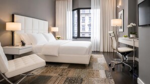 A modern hotel room with a large bed, a desk, a chair, and a view of a building through the window.