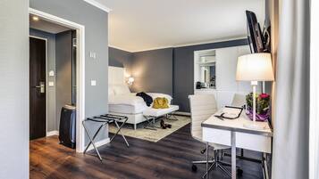 A modern hotel room with a bed, a desk with a lamp, a chair, a small table, and a suitcase.