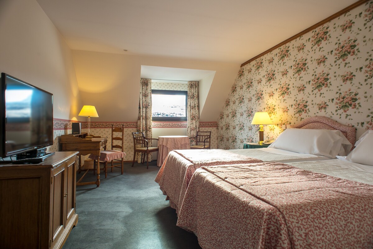 Hotelkamer met televisie, bureau met lamp, stoel, klein tafeltje en bed met bloemenbehang.