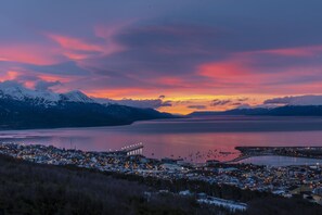 A coastal town at dusk with a bridge, mountains, and a calm sea.