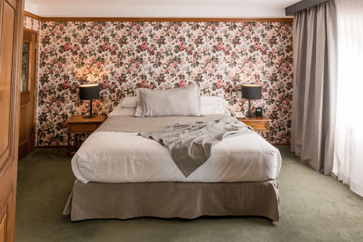 Un lit bien fait, agrémenté d’un couvre-lit gris, de deux lampes de chevet et d’un papier peint à motifs floraux.