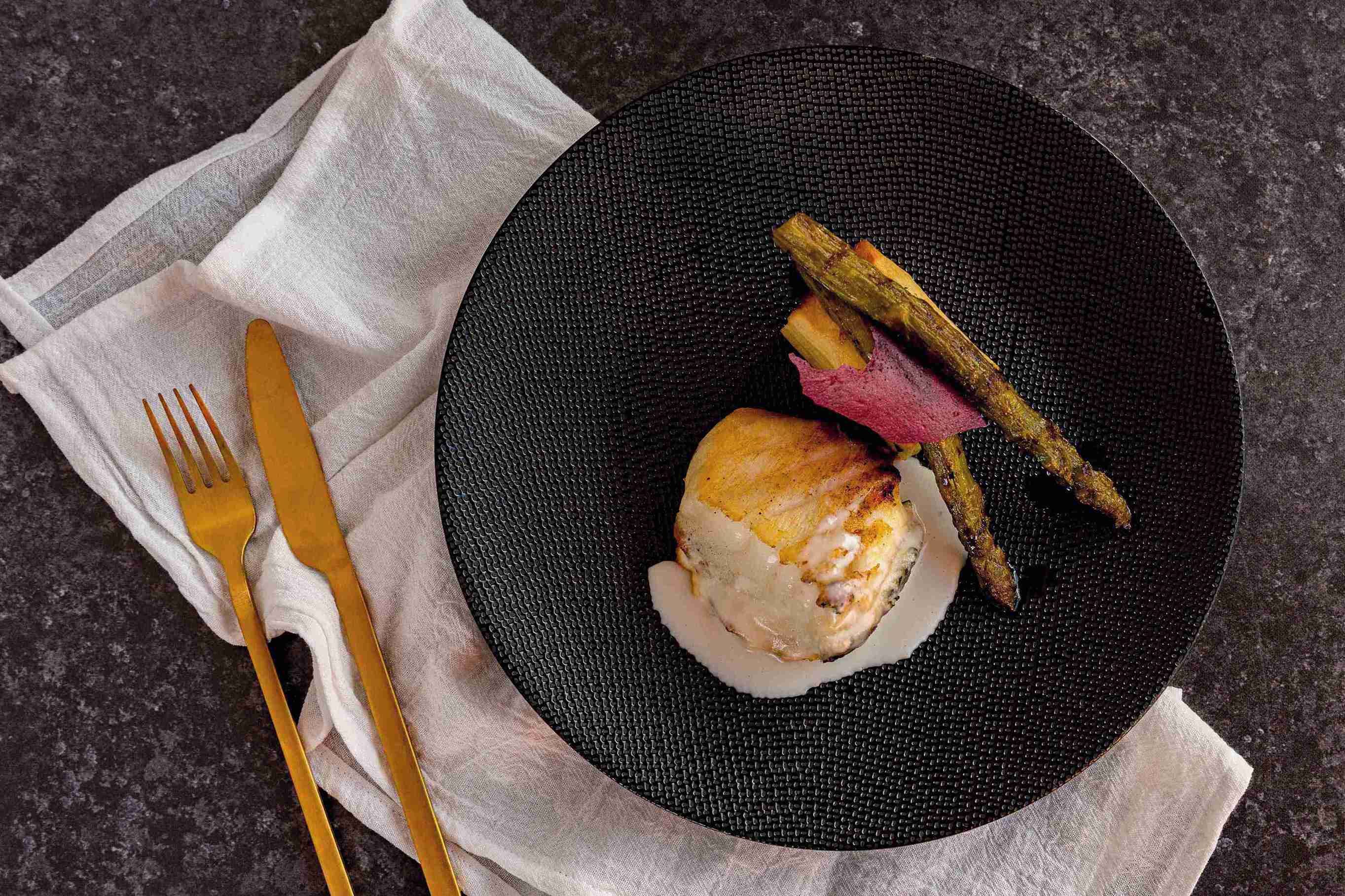 A plate with a piece of meat and vegetables on a dark textured plate, accompanied by a white napkin and a gold fork and knife.