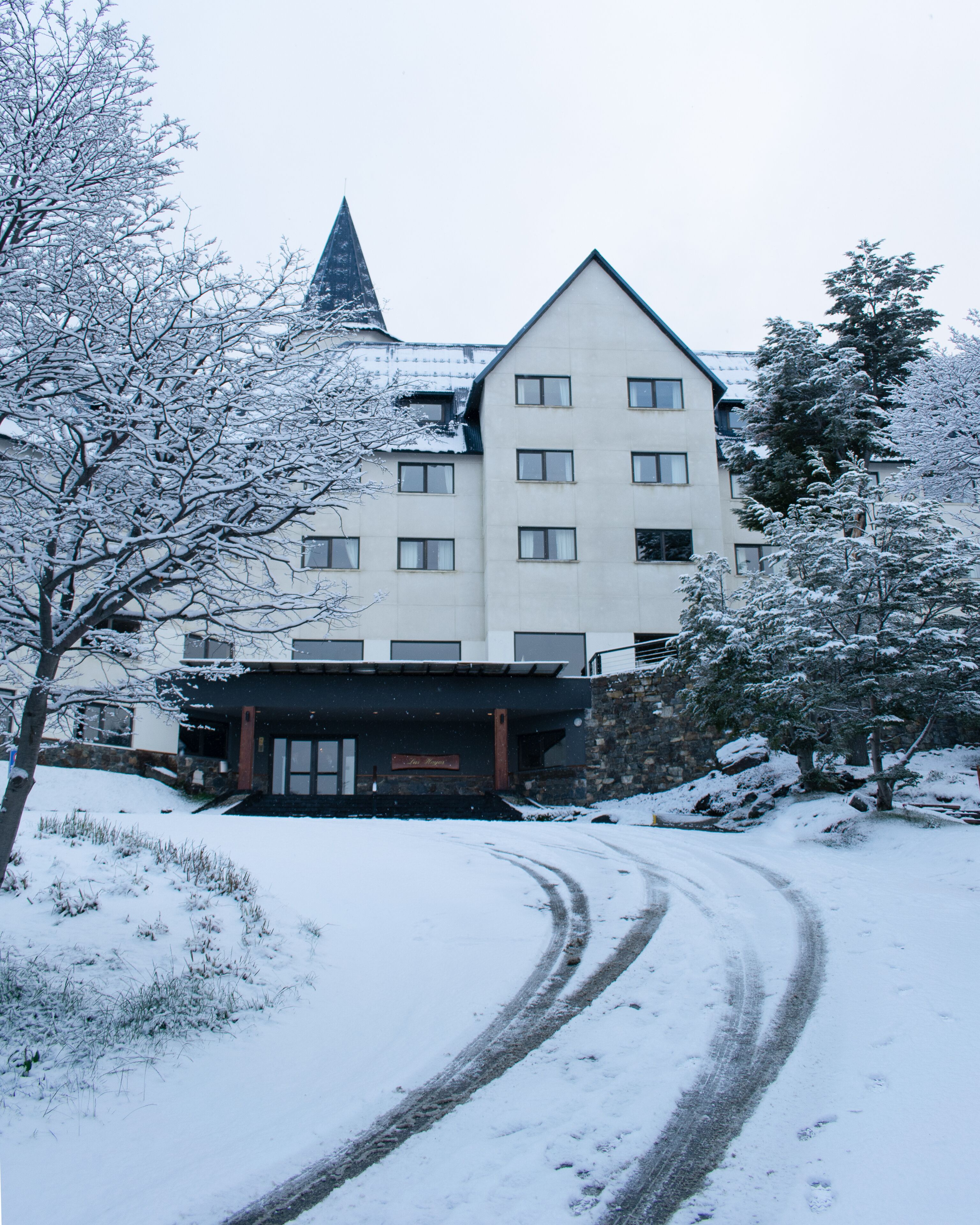 Un edificio cubierto de nieve con una torre destacada, un camino pavimentado y árboles cargados de nieve.