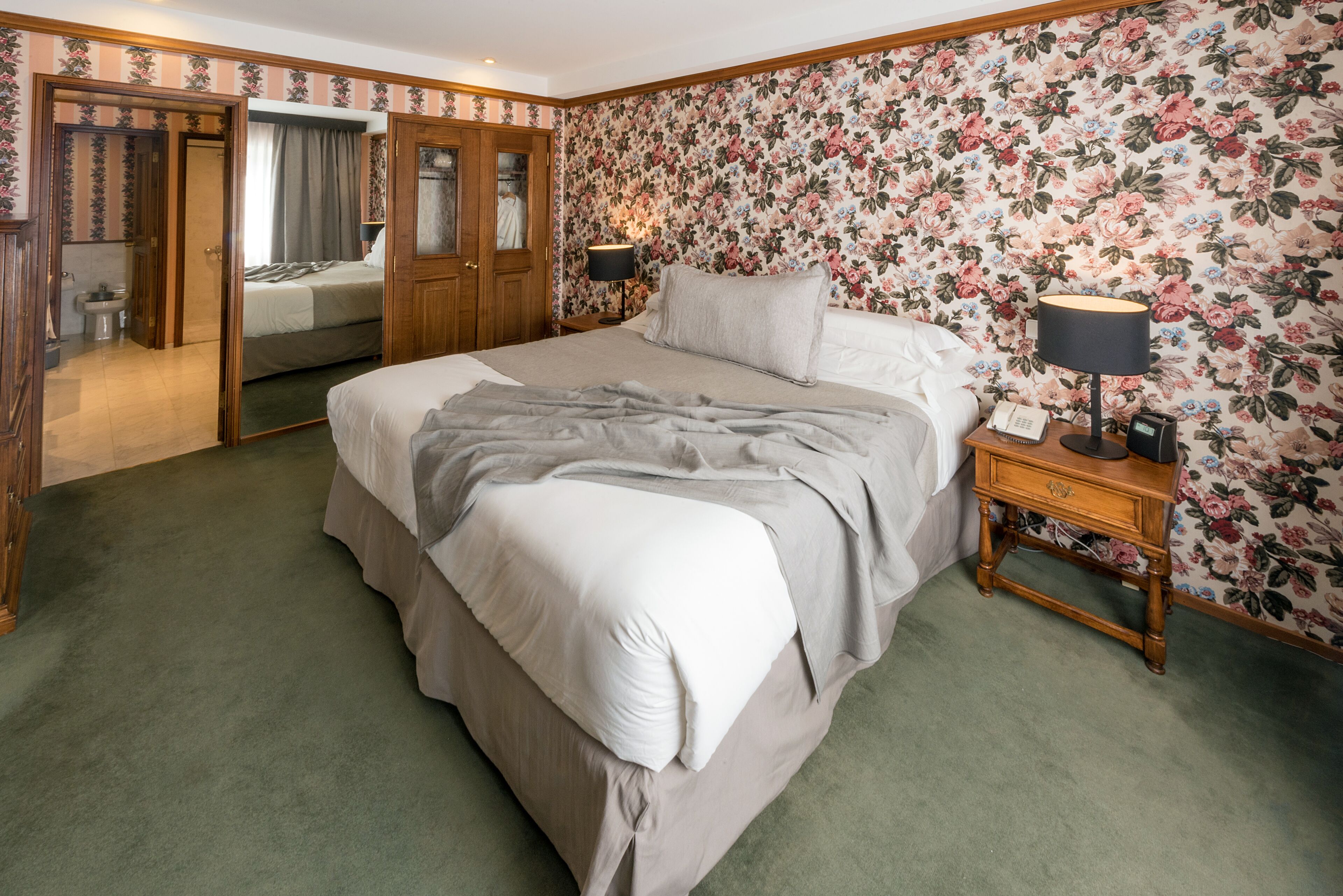 A bedroom with a large bed, a nightstand with a lamp, a wooden wardrobe, and a floral wallpaper.