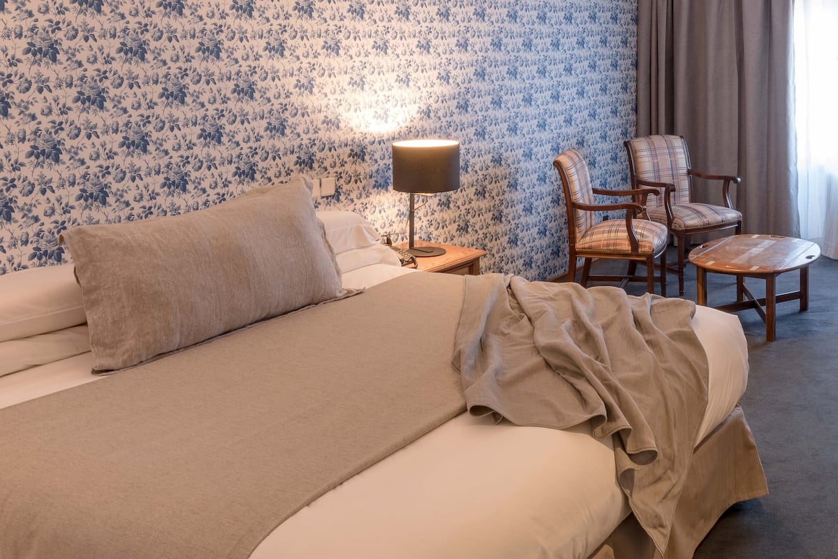 Un lit bien fait, recouvert d’une couverture beige, une lampe de chevet et deux fauteuils à motifs écossais dans une pièce dont le papier peint est à fleurs.
