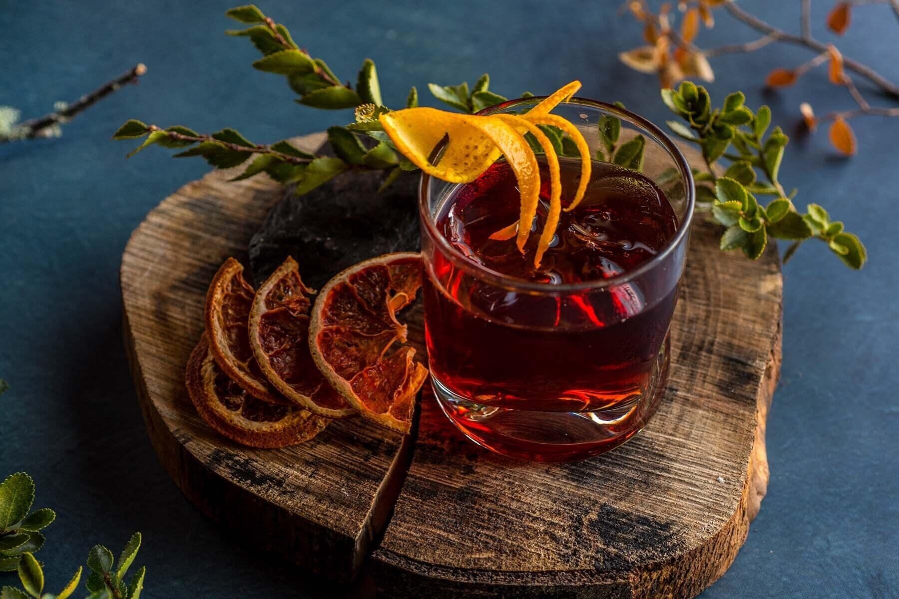 A glass of red beverage with dried orange slices and a garnish, placed on a wooden board with dried orange slices and green leaves around it.