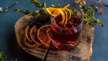 A glass of red beverage with dried orange slices and a garnish, placed on a wooden board with dried orange slices and green leaves around it.