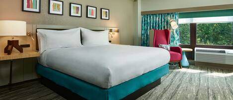 A hotel room with a large bed, a desk with a lamp, a chair, and a window with a view of trees.