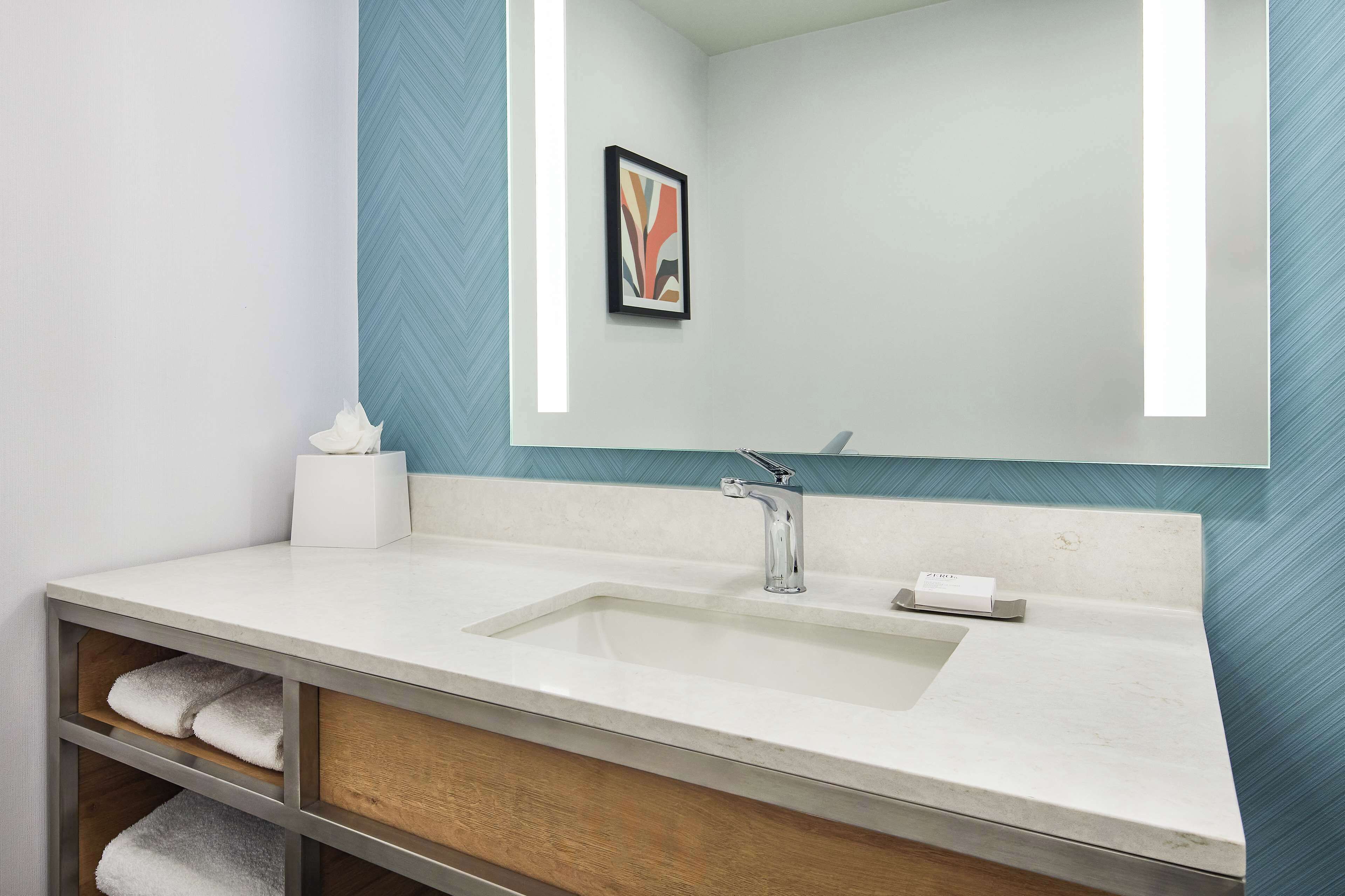 Un baño con un lavabo blanco, un espejo, toallero y un cuadro en la pared.