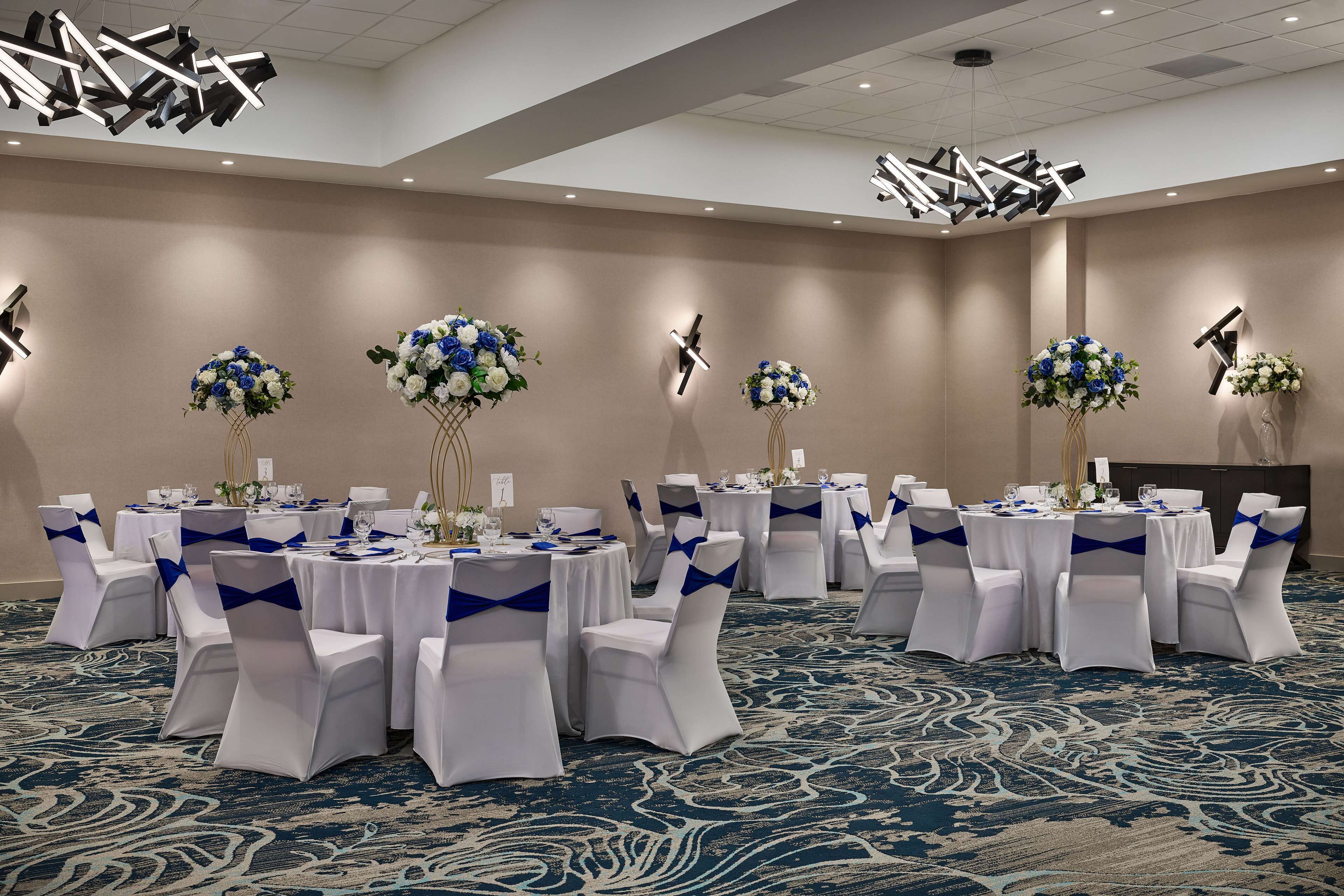 A banquet hall with round tables, white chairs with blue sashes, and floral centerpieces.
