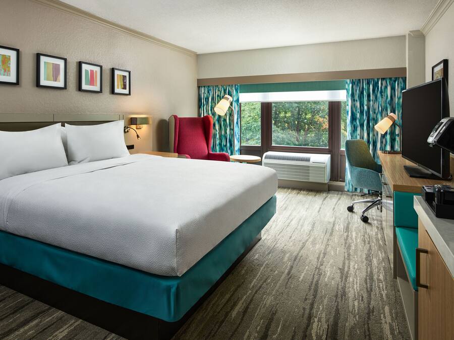A hotel room with a large bed, a desk with a computer, a TV, and a window with curtains.
