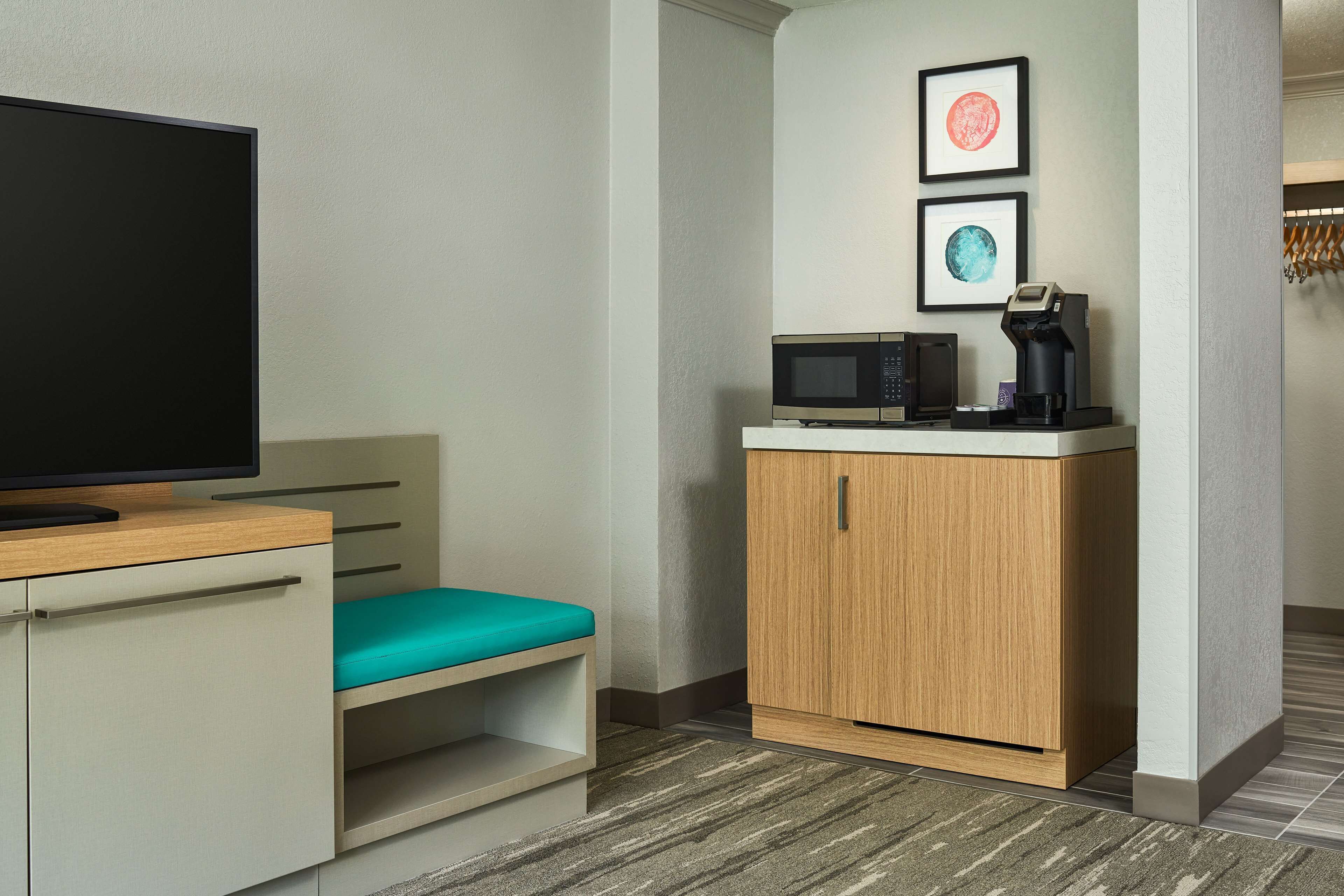 Ein Hotelzimmer mit Fernseher, Mikrowelle, Kaffeemaschine, einer türkisblauen Bank und einem Holzkabinent.