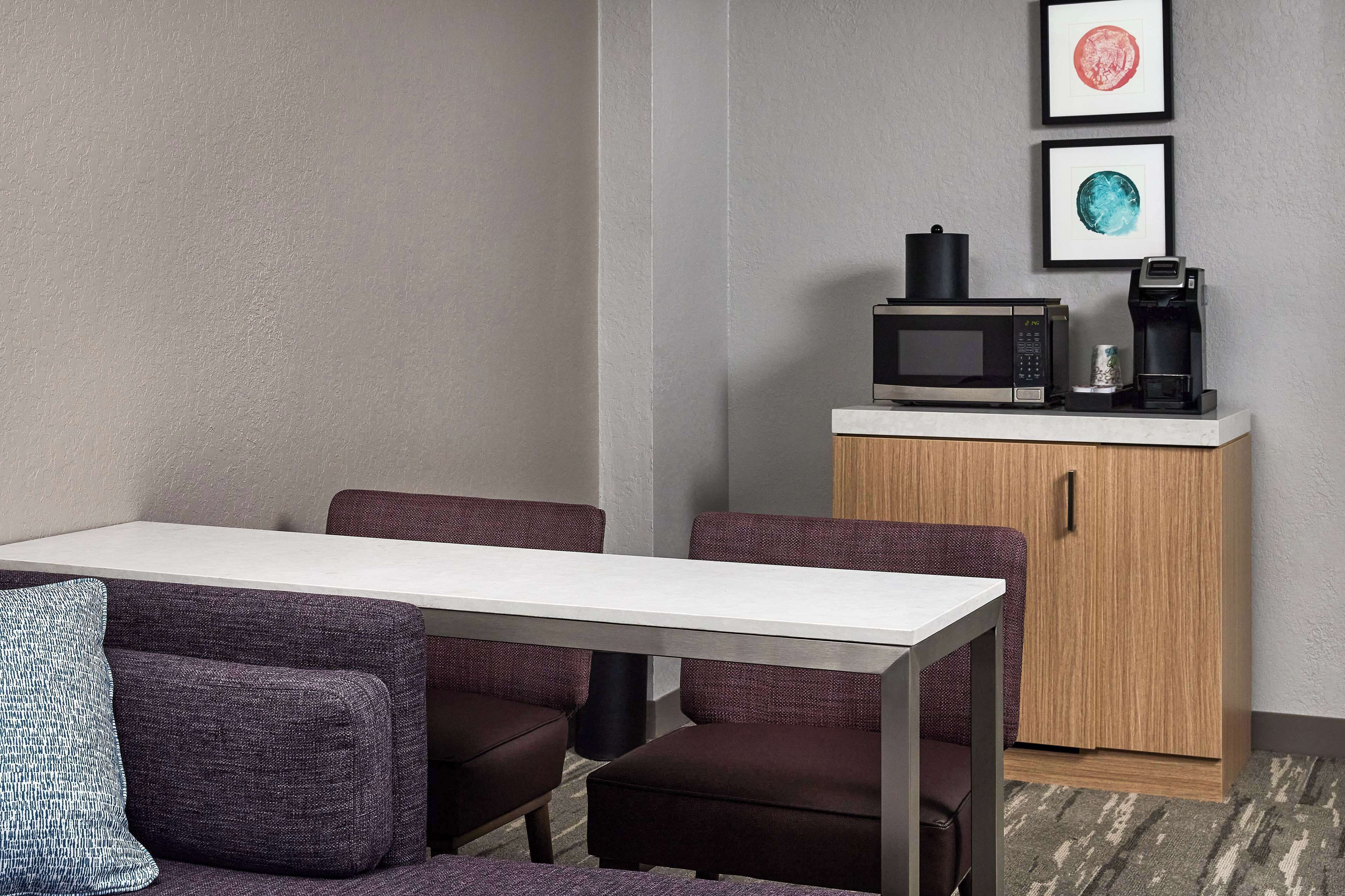 Una habitación de hotel moderna con zona de comedor que incluye una mesa blanca y sillas tapizadas de color púrpura. Un microondas y una cafetera están sobre un mueble de madera, y dos cuadros enmarcados decoran la pared.