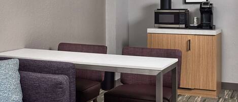 A modern hotel room with a dining area featuring a white table and purple upholstered chairs. A microwave and coffee maker are on a wooden cabinet, and two framed pictures are on the wall.