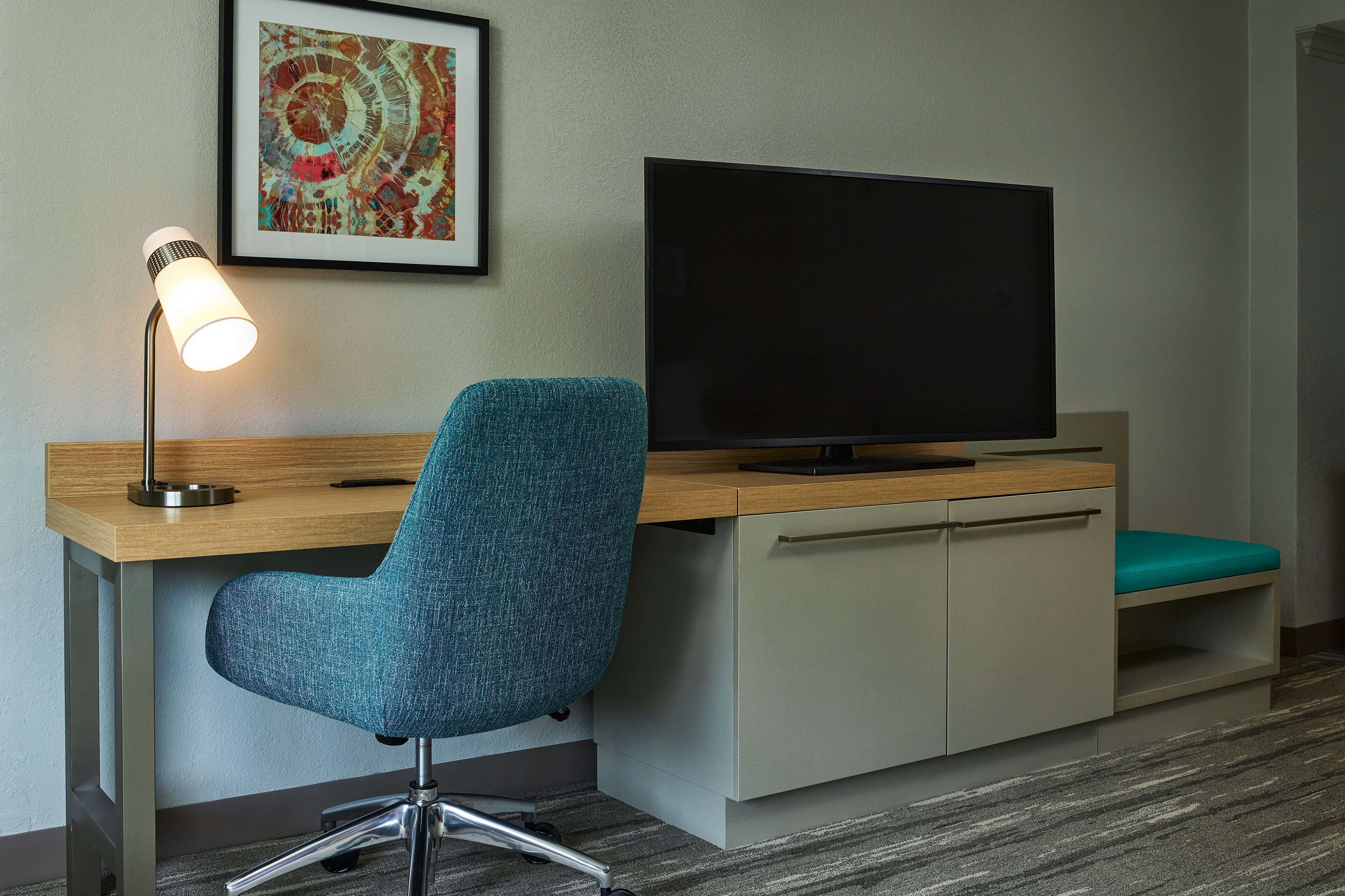 A hotel room with a desk, a teal office chair, a flat-screen TV, a framed artwork, and a carpeted floor.