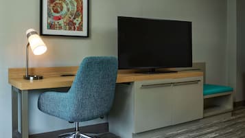 A hotel room with a desk, a teal office chair, a flat-screen TV, a framed artwork, and a carpeted floor.