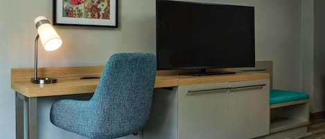 Habitación de hotel con escritorio, una silla de oficina color verde azulado, televisor de pantalla plana, una obra de arte enmarcada y piso cubierto de alfombra.