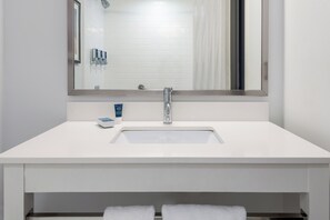 Une salle de bain moderne avec un lavabo blanc, un grand miroir et une douche visible au fond.