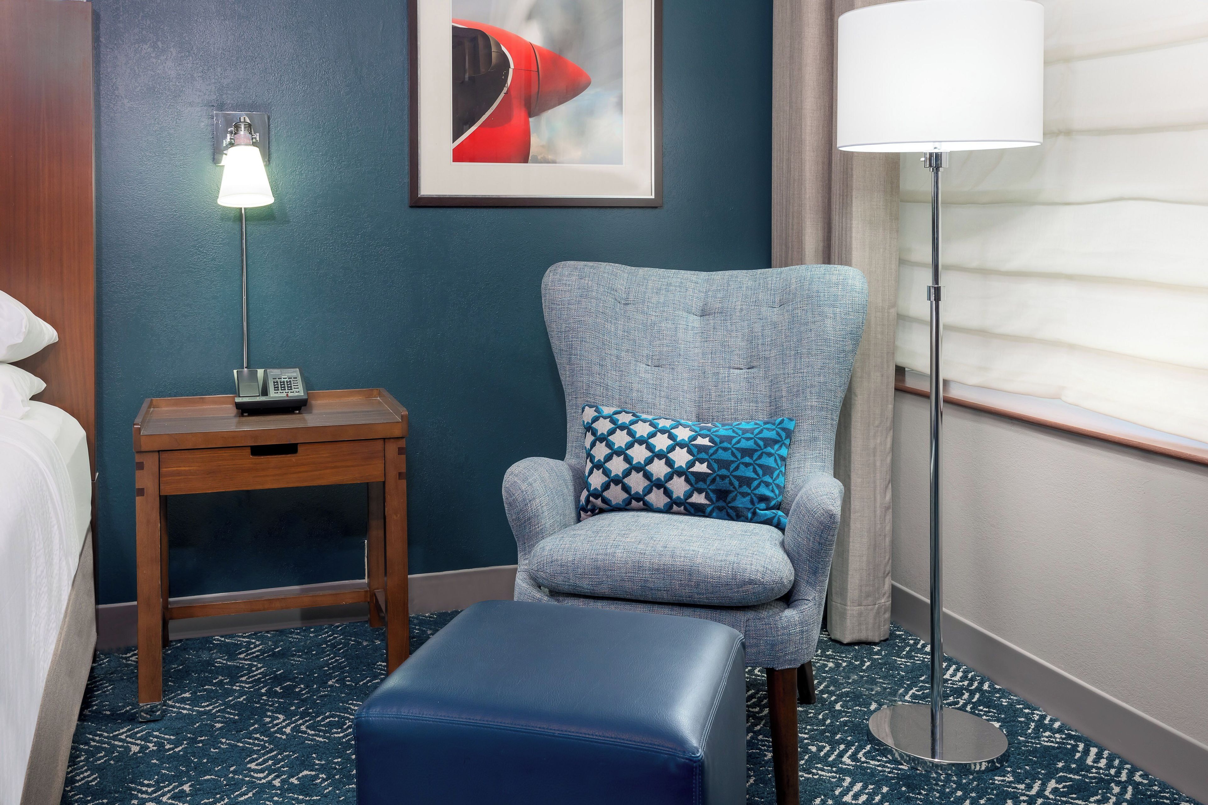 A hotel room with a blue armchair, a small table with a lamp, a framed picture on the wall, and a large window with curtains.