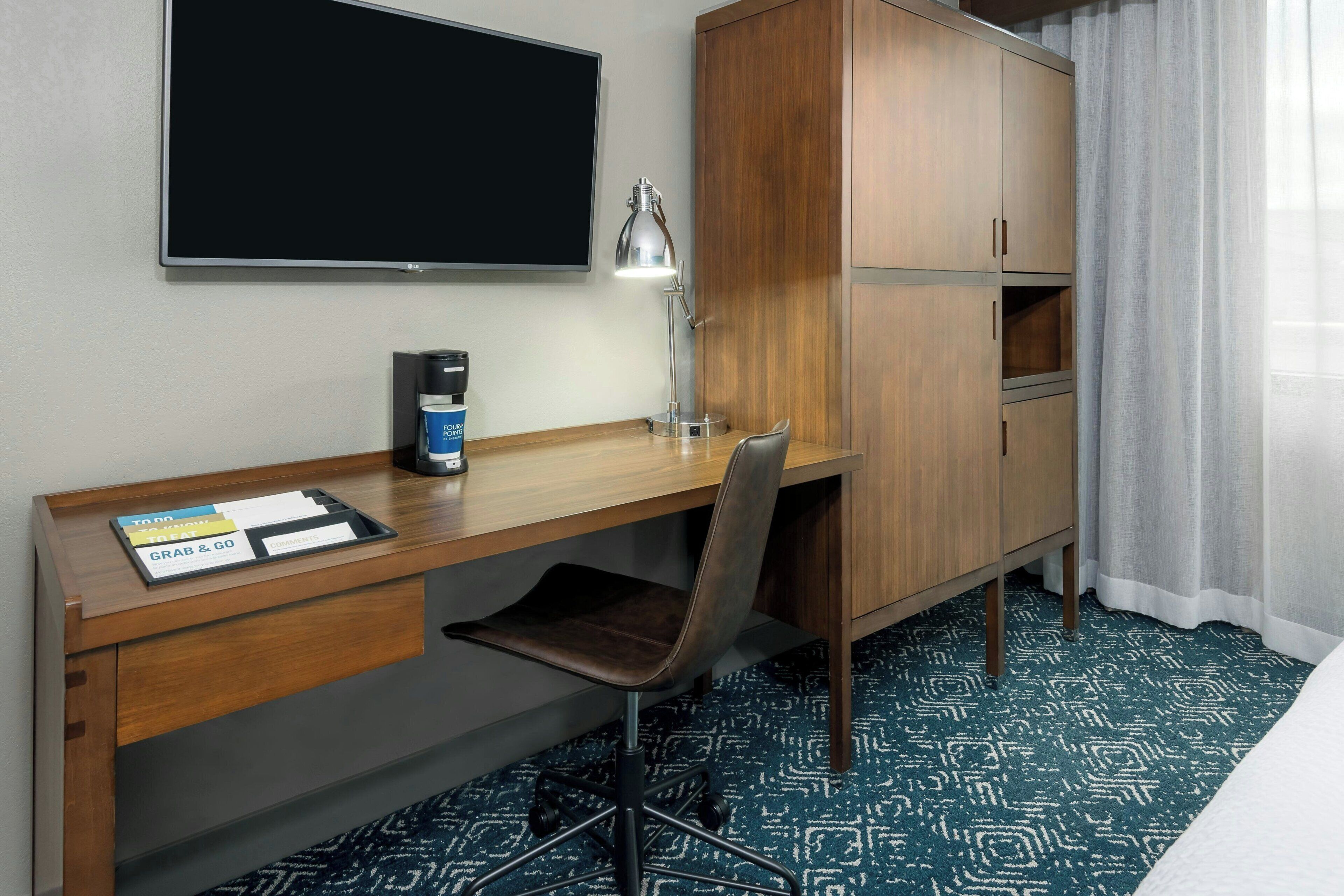 A hotel room with a wooden desk, a chair, a flat-screen TV, a lamp, a coffee maker, and a wardrobe.