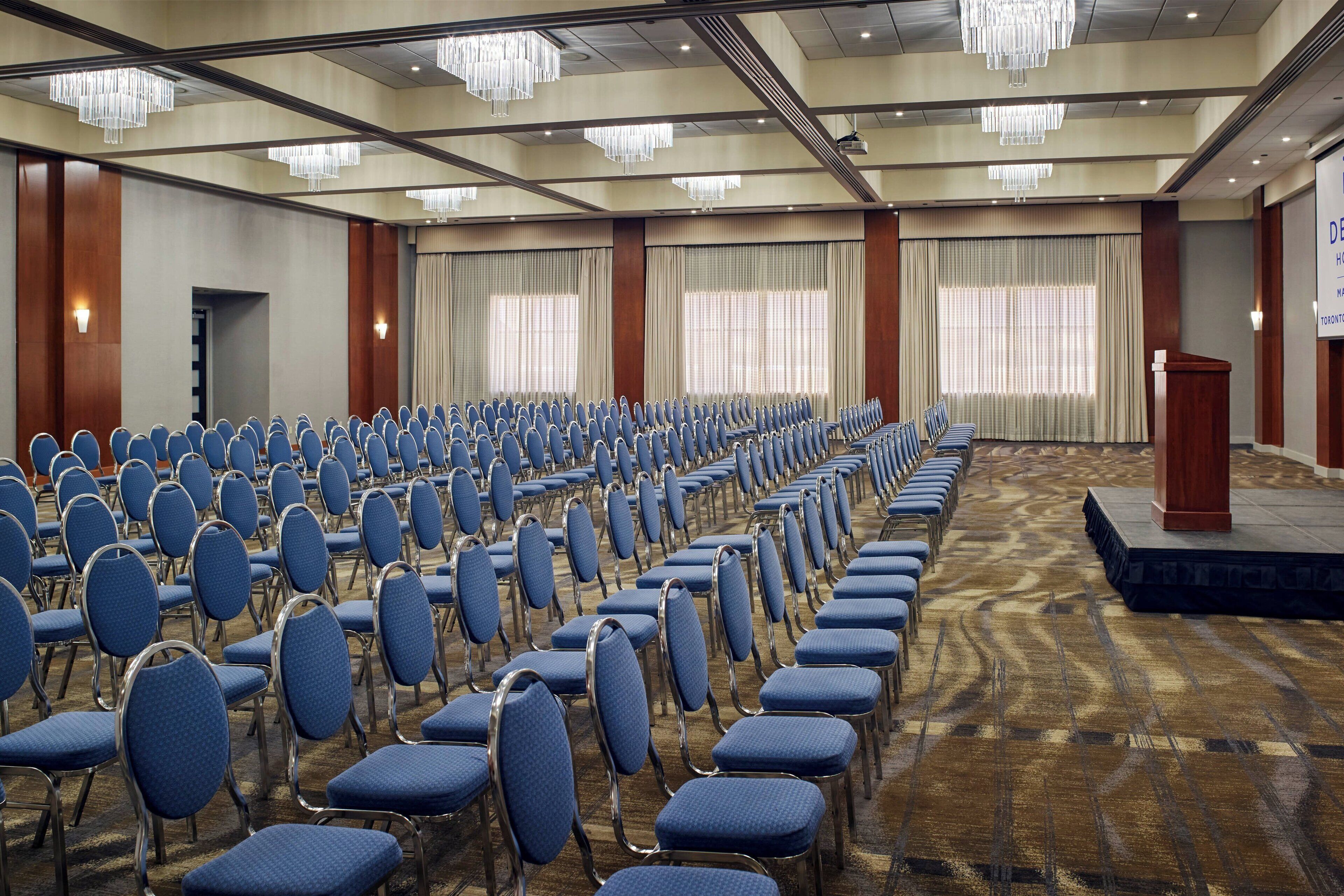 A conference room with rows of blue chairs, a podium, and large windows with curtains.