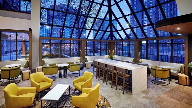 A spacious lounge area with yellow armchairs, a central coffee table, and a long bar. Large windows offer views of the cityscape. - Delta Hotels by Marriott Toronto Mississauga (Mississauga)