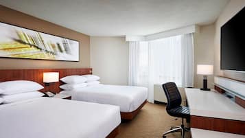 Habitación de hotel con dos camas, un escritorio, una silla, un televisor y un ventanal grande con cortinas.