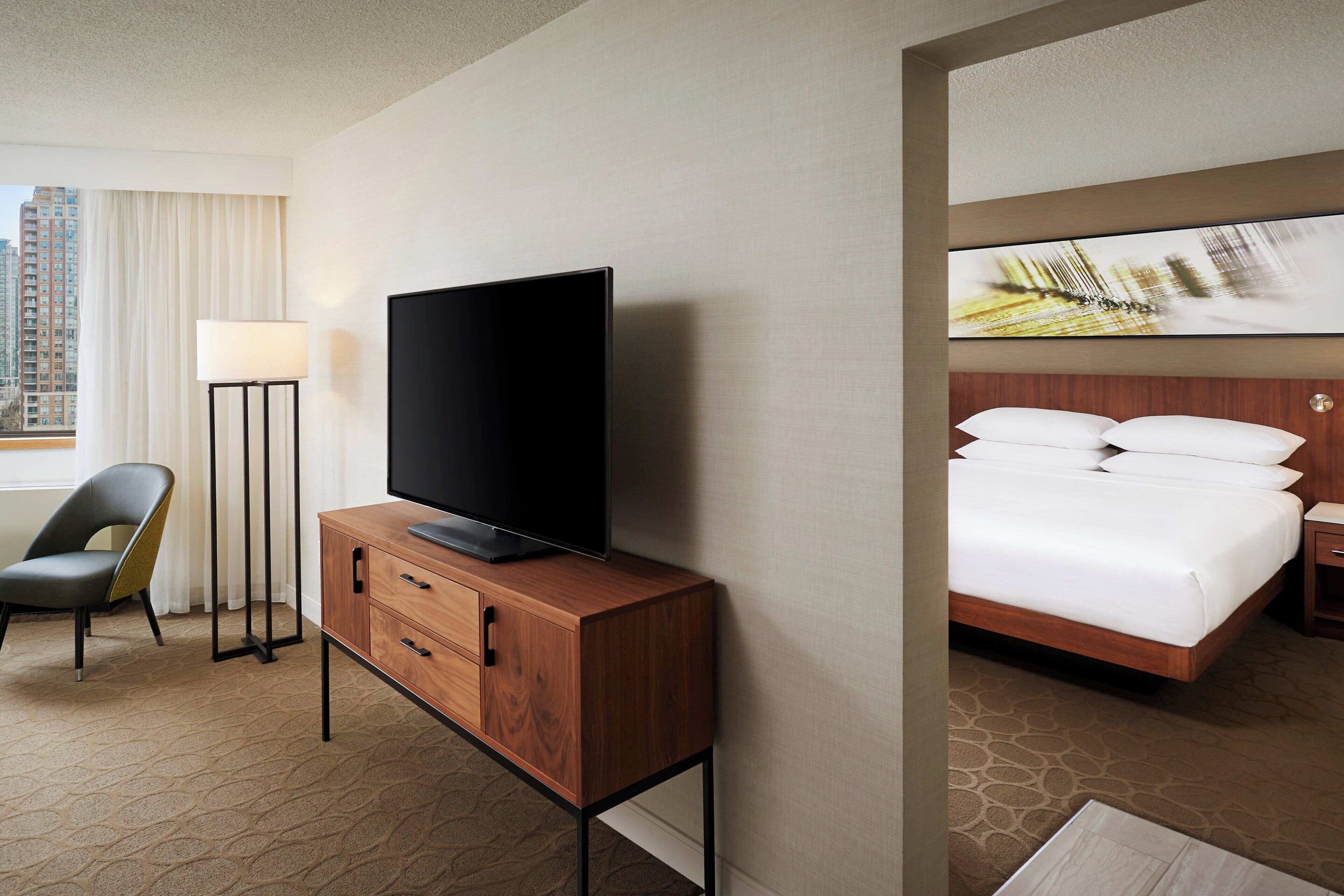 Habitación de hotel con televisión sobre un mueble de madera, una cama con ropa blanca y vistas a edificios a través del ventanal.