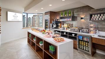 Zona de desayuno de hotel con un sistema de buffet, que incluye un refrigerador con bebidas, una estación de café y diversos alimentos exhibidos en una larga mesa.