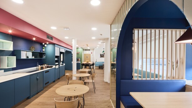 Un interior moderno con área de cocina, mesas de comedor y zona de estar con pared azul.