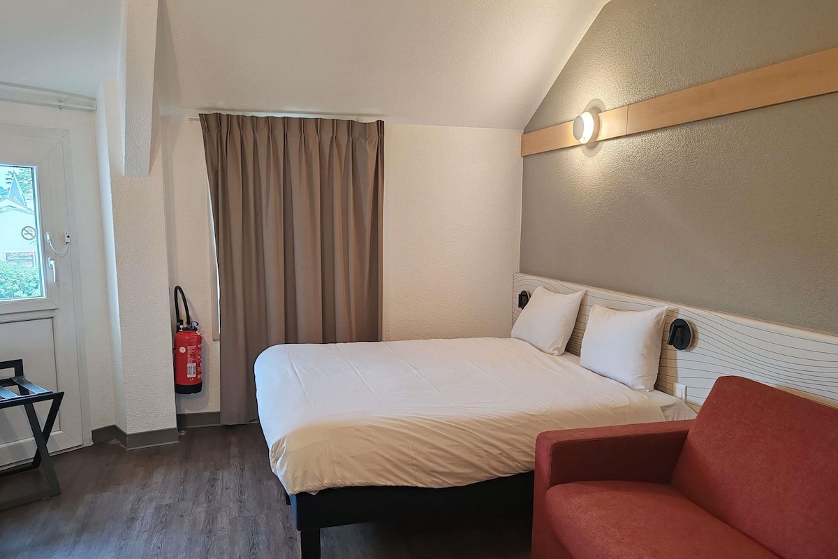 Une chambre d’hôtel avec un lit, un fauteuil rouge, un extincteur et une fenêtre avec des rideaux.