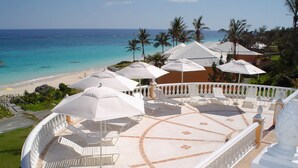 A resort balcony with white lounge chairs and umbrellas, overlooking a beach and ocean.