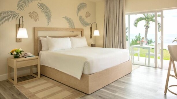 A hotel room with a large bed, a nightstand with a lamp, a view of the outdoors through a sliding door, and palm trees visible outside.