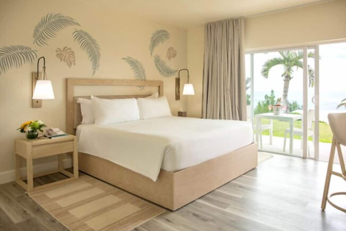 A hotel room with a large bed, a nightstand with a lamp, a view of the outdoors through a sliding door, and palm trees visible outside.