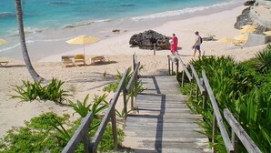 A wooden boardwalk leading to a beach with clear turquoise water, palm trees, and beach umbrellas.