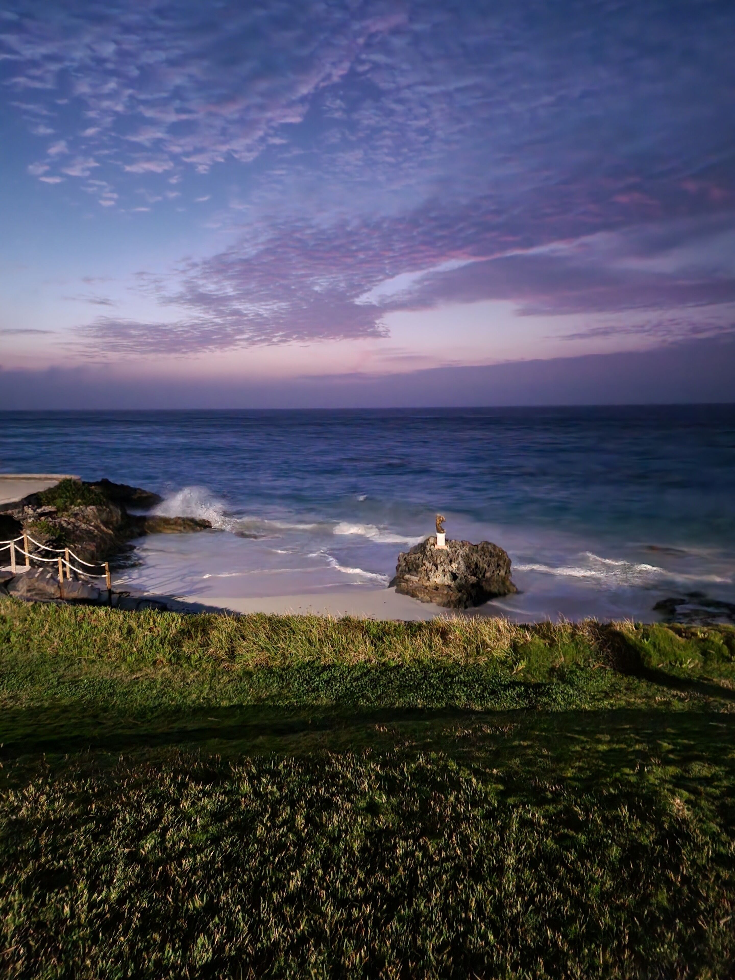 A coastal landscape with a rocky outcrop and a statue, a grassy foreground, and a view of the ocean.