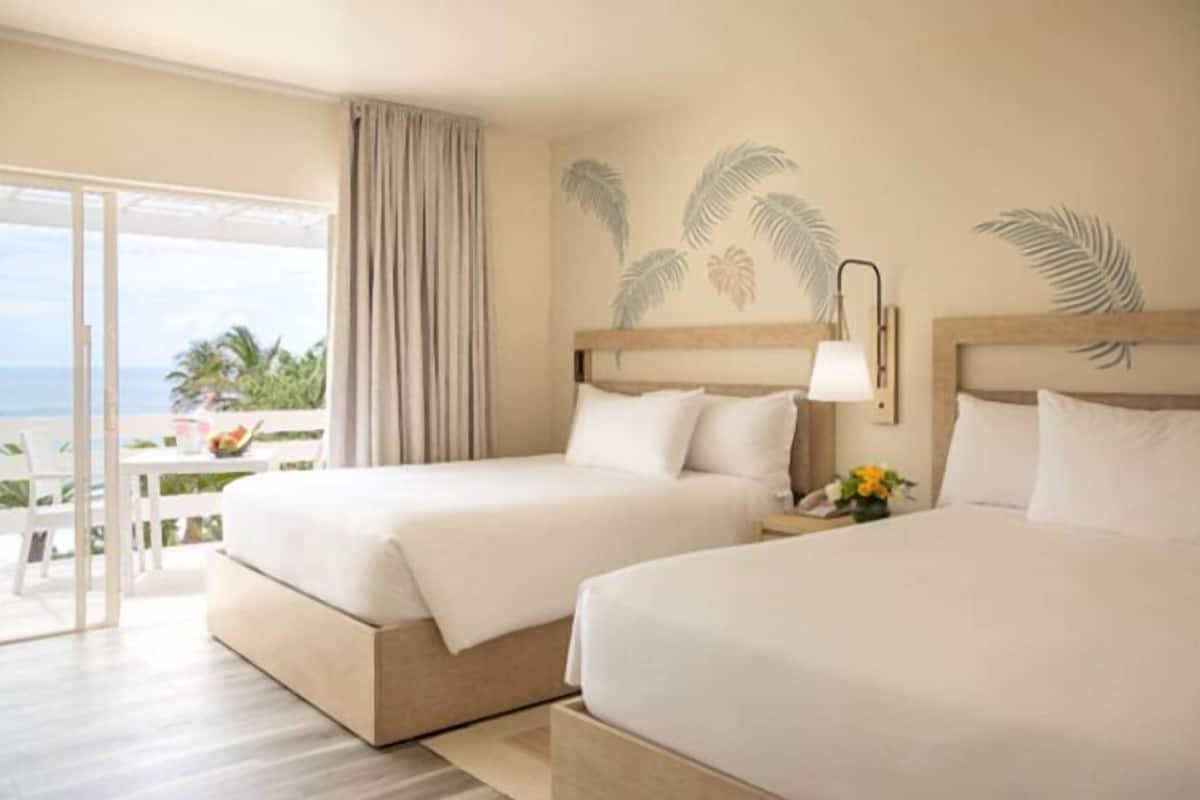 A hotel room with two beds, a balcony with a view of the ocean, and a tropical wall design.