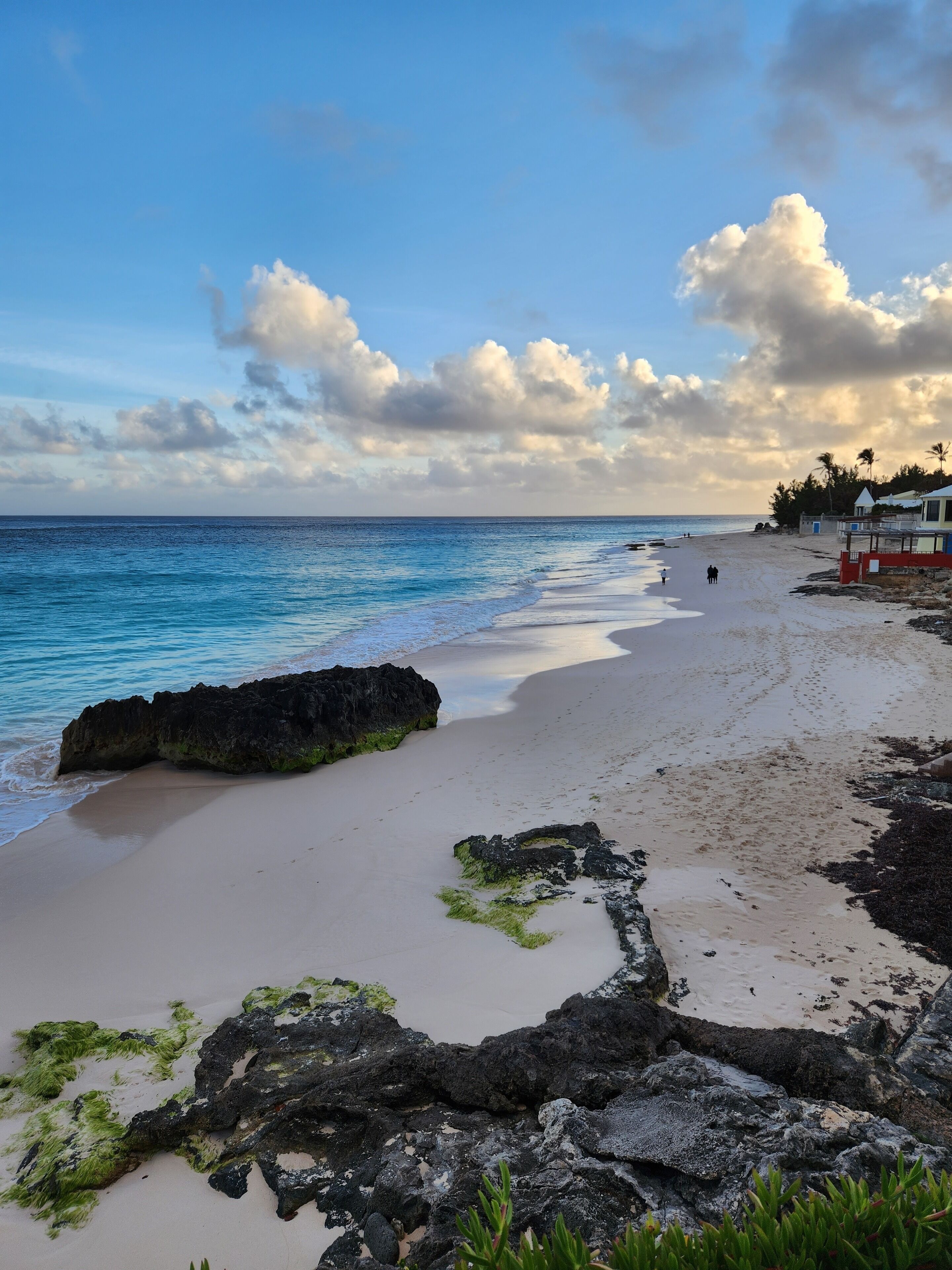 Ein Strand mit klarem, blauem Wasser, Felsen und ein paar Spaziergängern.