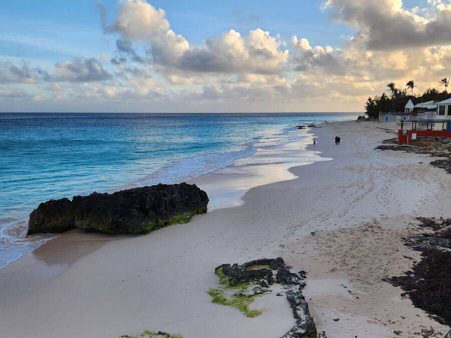 Una playa con agua azul cristalina, afloramientos rocosos y algunas personas caminando.