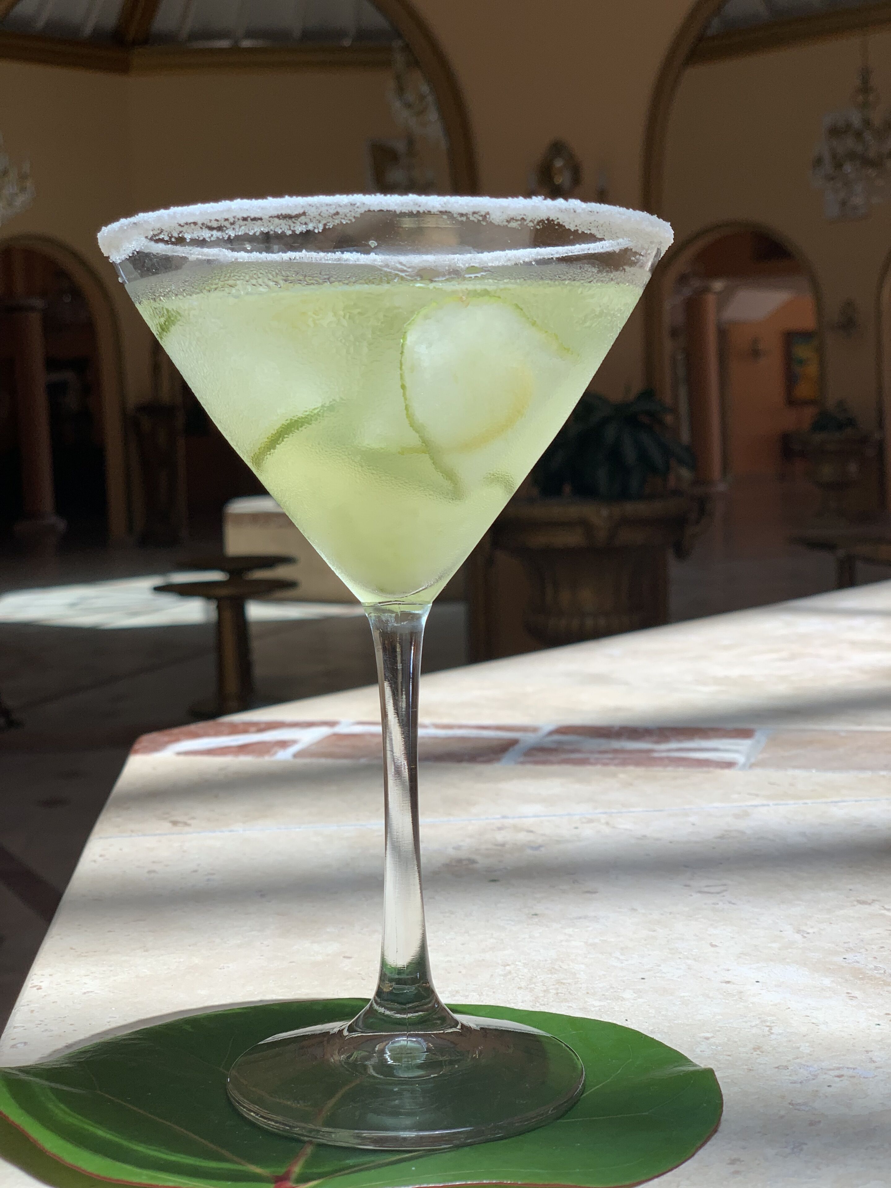 A martini glass with a salted rim on a green leaf, placed on a table in a room with arched ceilings and a chandelier.