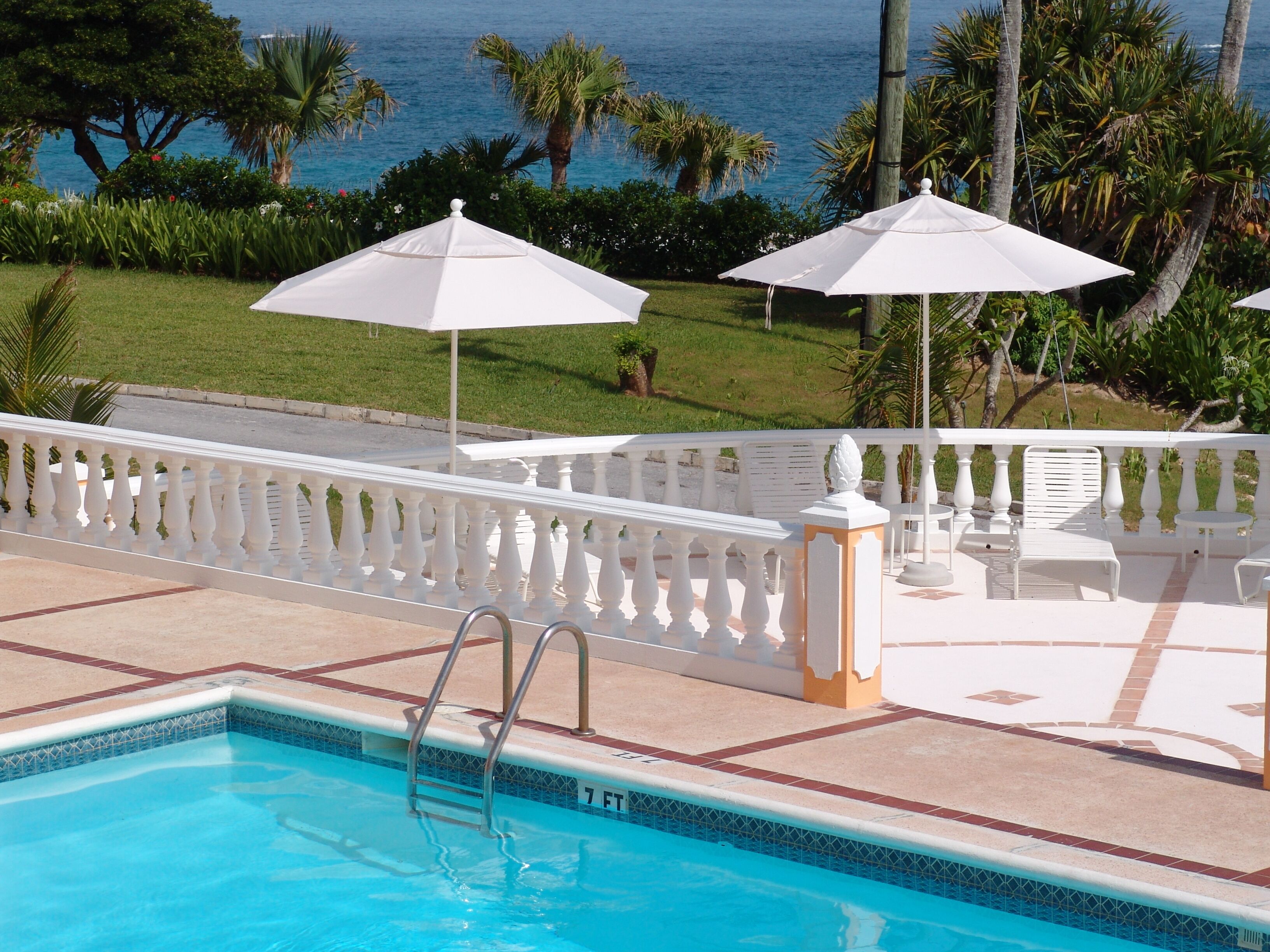 A swimming pool with a white fence, white umbrellas, and a view of the ocean.