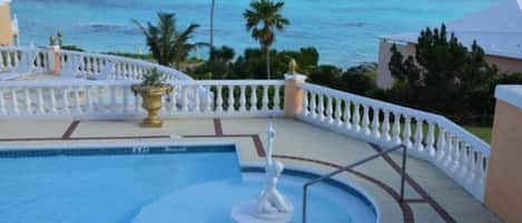 A pool with a fountain, surrounded by a white balustrade, overlooking a beach with palm trees and a clear view of the ocean.